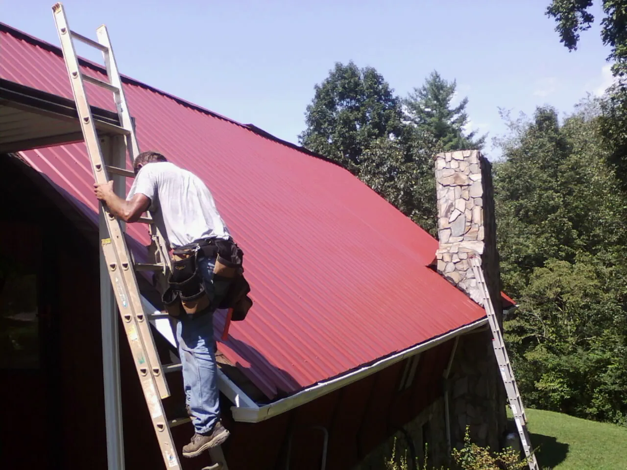 Roofer on a ladder beside a red metal roof and stone chimney — Soffit & Fascia Repair by Roofing Brothers in Wilkesboro